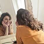 A cheerful woman smiles at her reflection in a vintage-style mirror, exuding positivity and warmth.